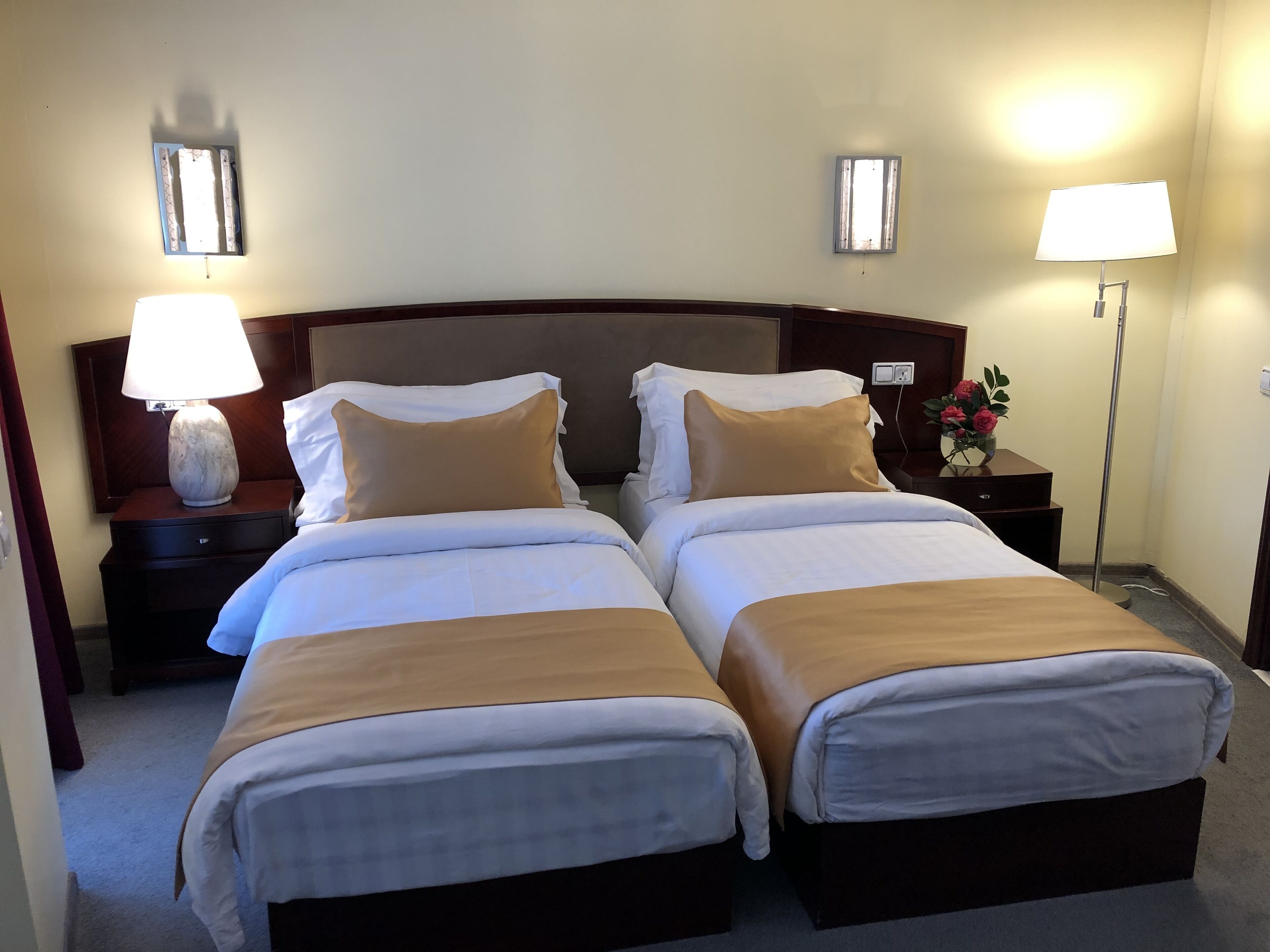Una habitació d'hotel amb dos llits, cadascun amb roba de llit blanca i beix, un capçal de fusta, làmpades de nit i una tauleta de nit amb un ram de flors.