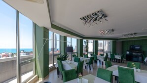 Un restaurante con sillas verdes y mesas blancas, amplios ventanales con vista al mar y un diseño de techo moderno.