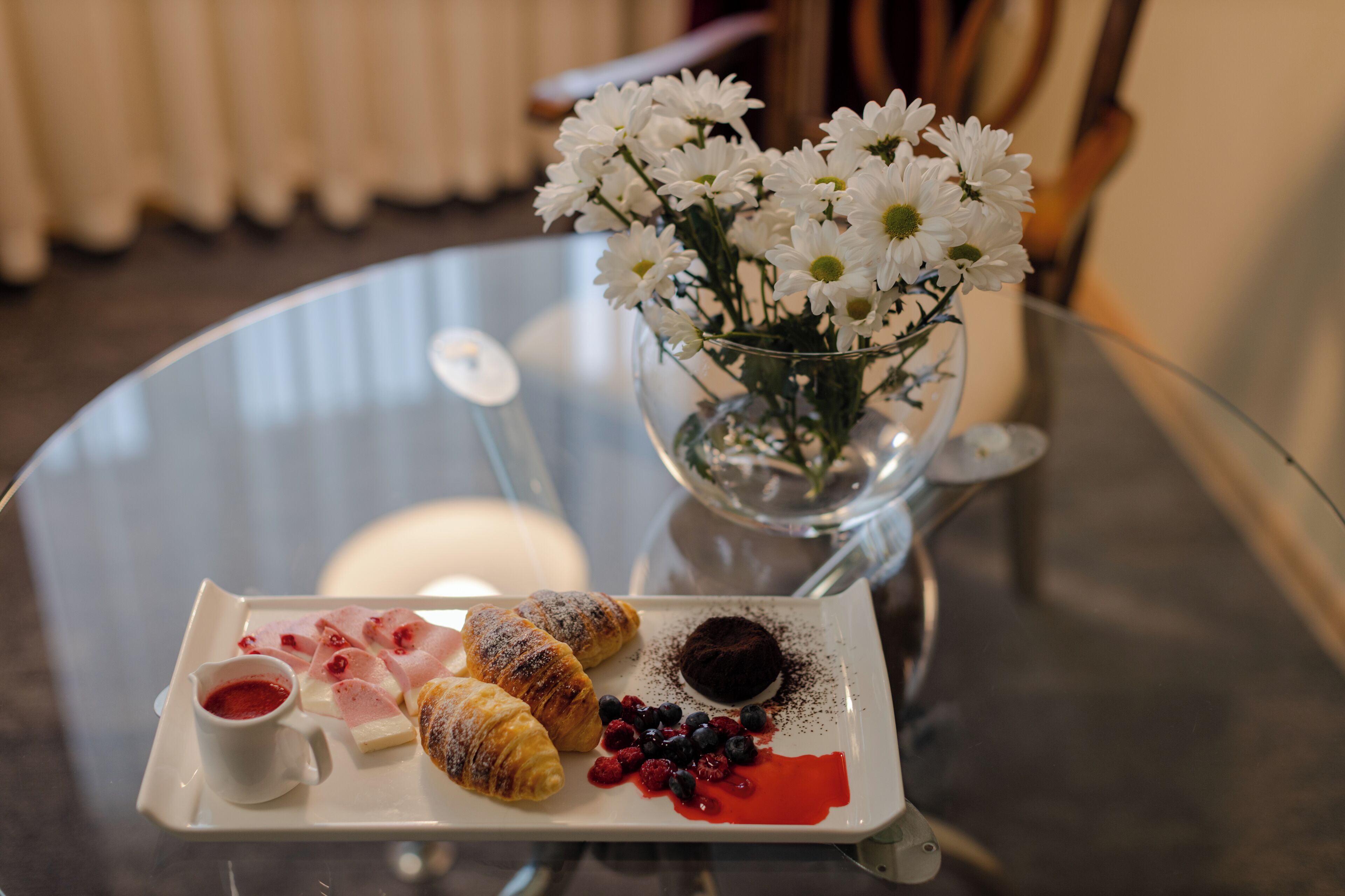 A glass table with a white plate of pastries, a small vase with white flowers, and a glass of red liquid.