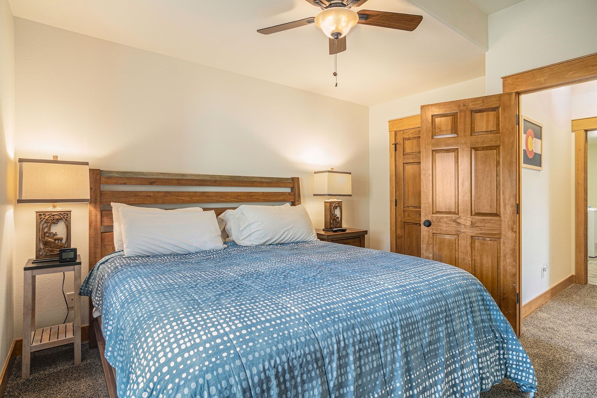 A bedroom with a wooden headboard, a bed with a blue polka dot bedspread, two bedside lamps, a nightstand, and a wooden door.