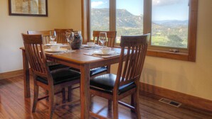 A dining room with a table set for four, wooden chairs, a vase, and a view of mountains through a large window.