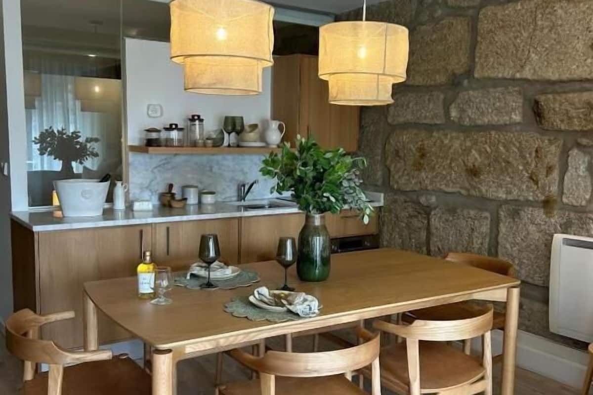A dining area with a wooden table and chairs, a vase with greenery, and pendant lights.