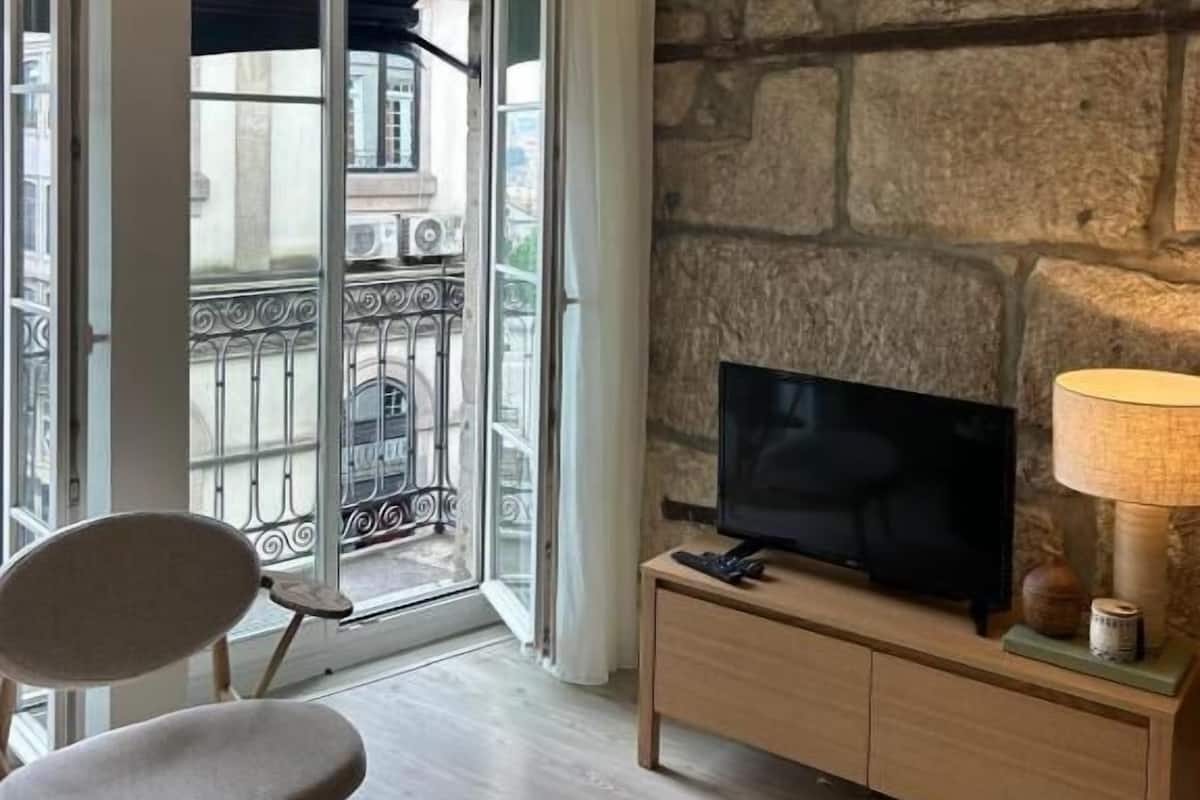 A living room with a stone wall, a television on a wooden stand, and a view of a balcony with a wrought iron railing.