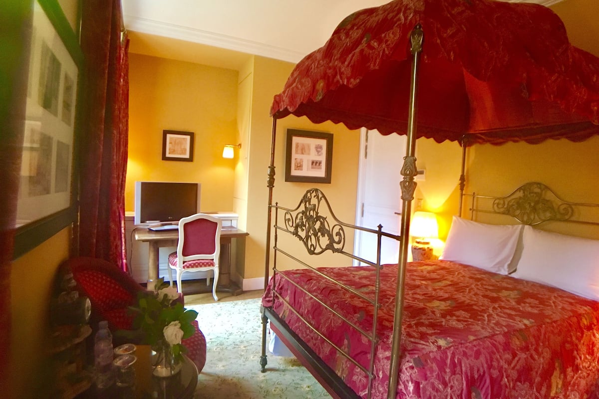A four-poster bed with a red canopy, a desk with a chair, and a television in a room with yellow walls.