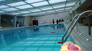 Une piscine intérieure entourée de parois en verre, avec un sol carrelé, une horloge au mur et un escalier menant à un niveau supérieur.