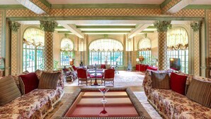 Un salon spacieux, doté de canapés à motifs floraux, d’une table basse centrale et de grandes fenêtres ornées de rideaux élégants.