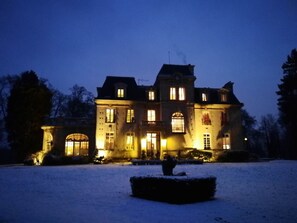 A large, illuminated mansion at night with multiple windows and a snow-covered lawn.