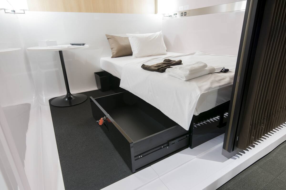 A modern hotel room with a bed, a bedside table, a small desk, and a storage unit under the bed.
