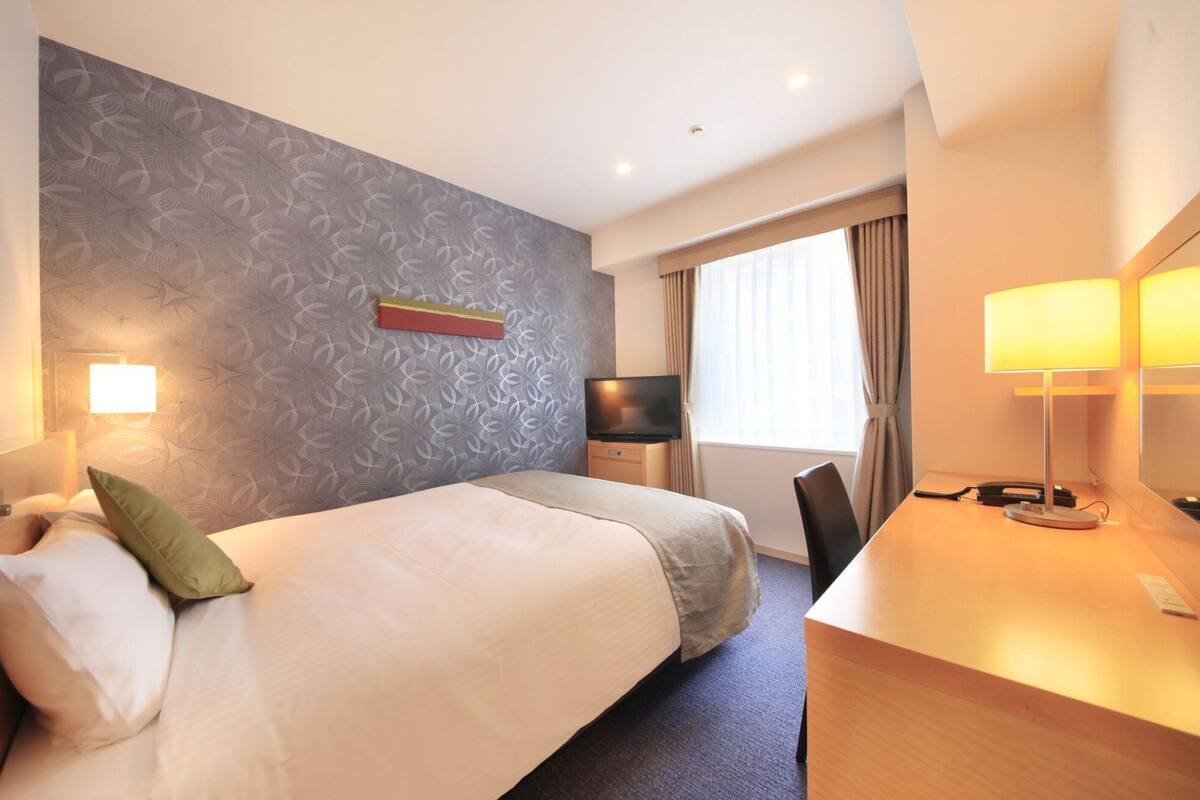 A hotel room with a bed, a desk with a lamp, a TV, and a window with curtains.