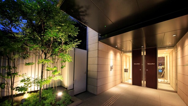 A modern building entrance with a gated driveway, a small garden area with trees, and a well-lit interior.