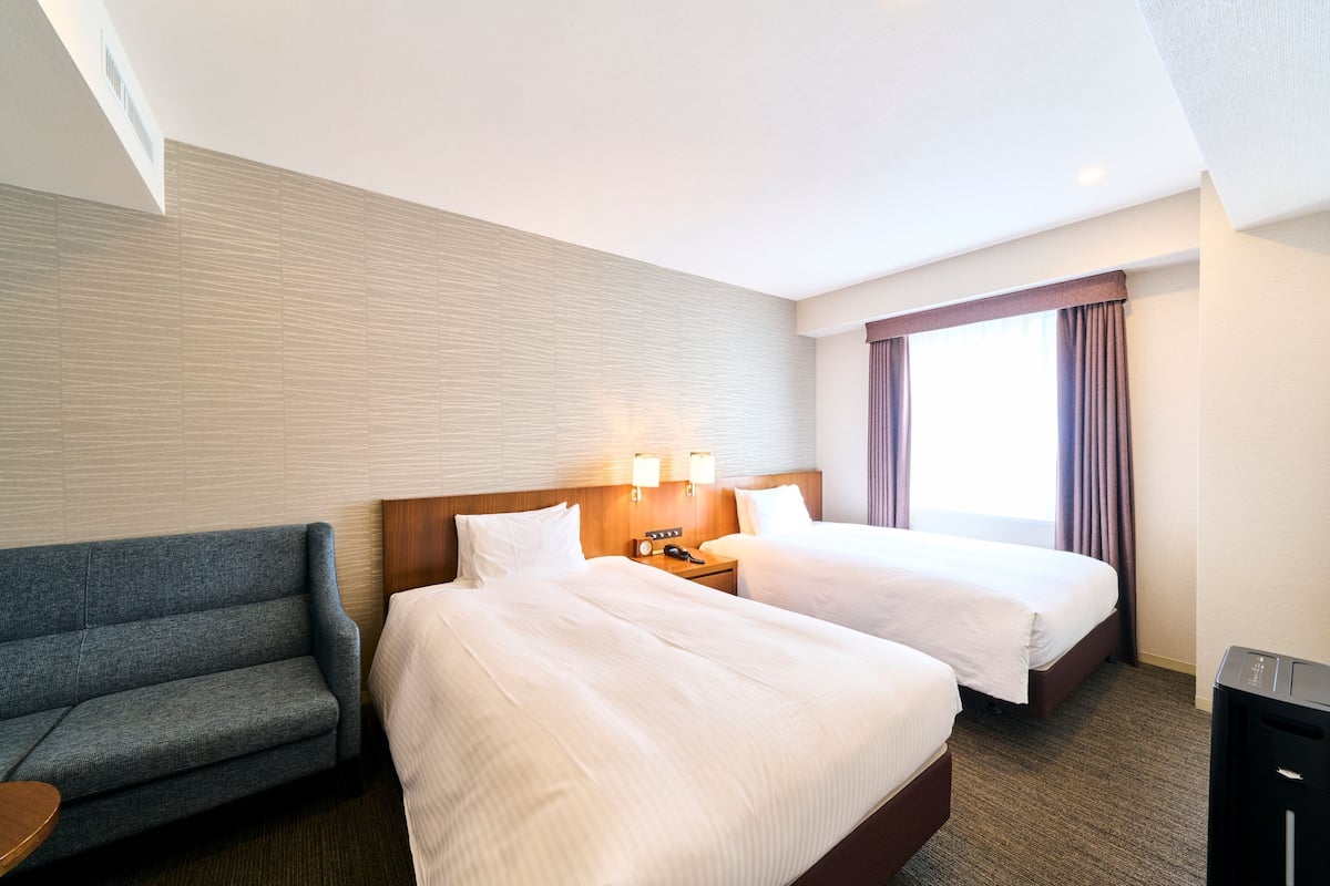 A hotel room with two beds, a sofa, a nightstand with a phone, and a window with curtains.