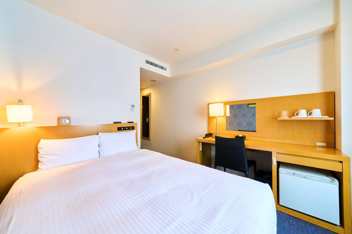 A hotel room with a bed, desk, chair, lamp, and a small refrigerator.