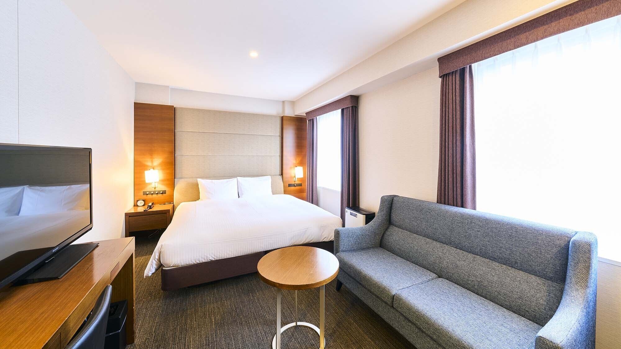 A hotel room with a bed, a TV, a sofa, a small table, and a nightstand with a lamp.
