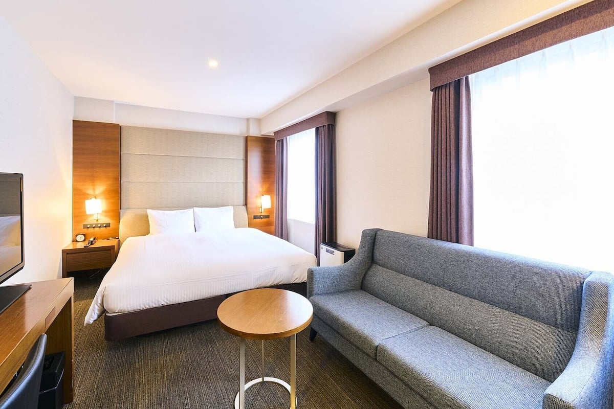 A hotel room with a bed, a TV, a sofa, a small table, and a nightstand with a lamp.