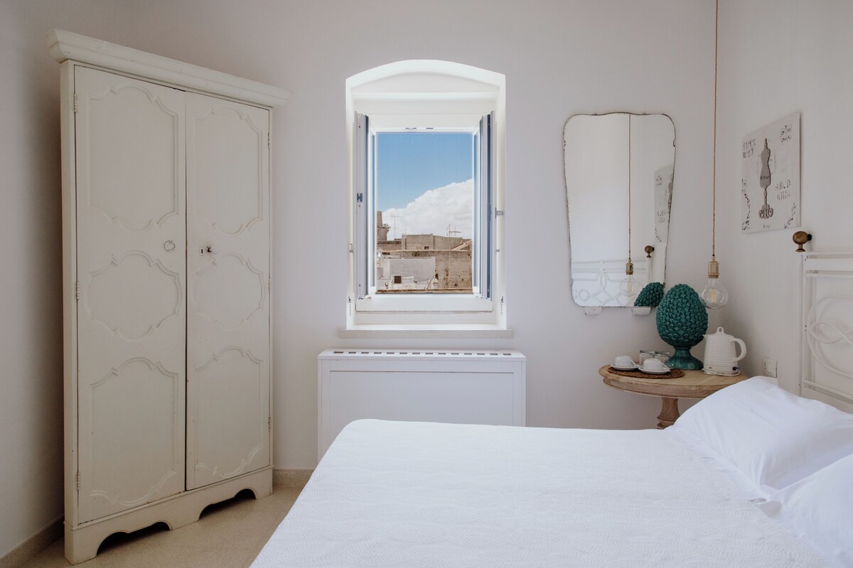 Un dormitorio con un armario grande, una mesita de noche con lámpara y una ventana con vistas a edificios.