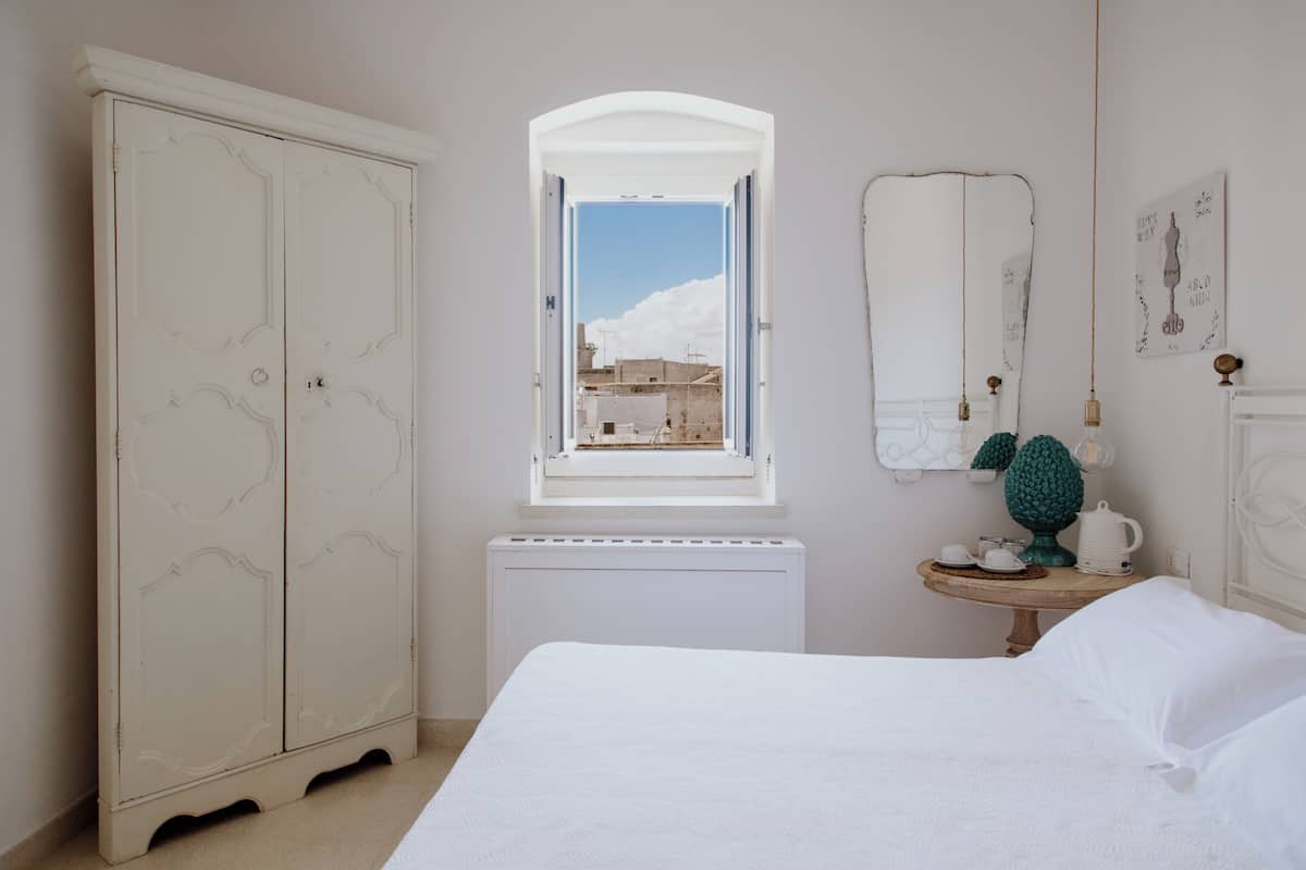 A bedroom with a large wardrobe, a small table with a lamp, and a window with a view of buildings.