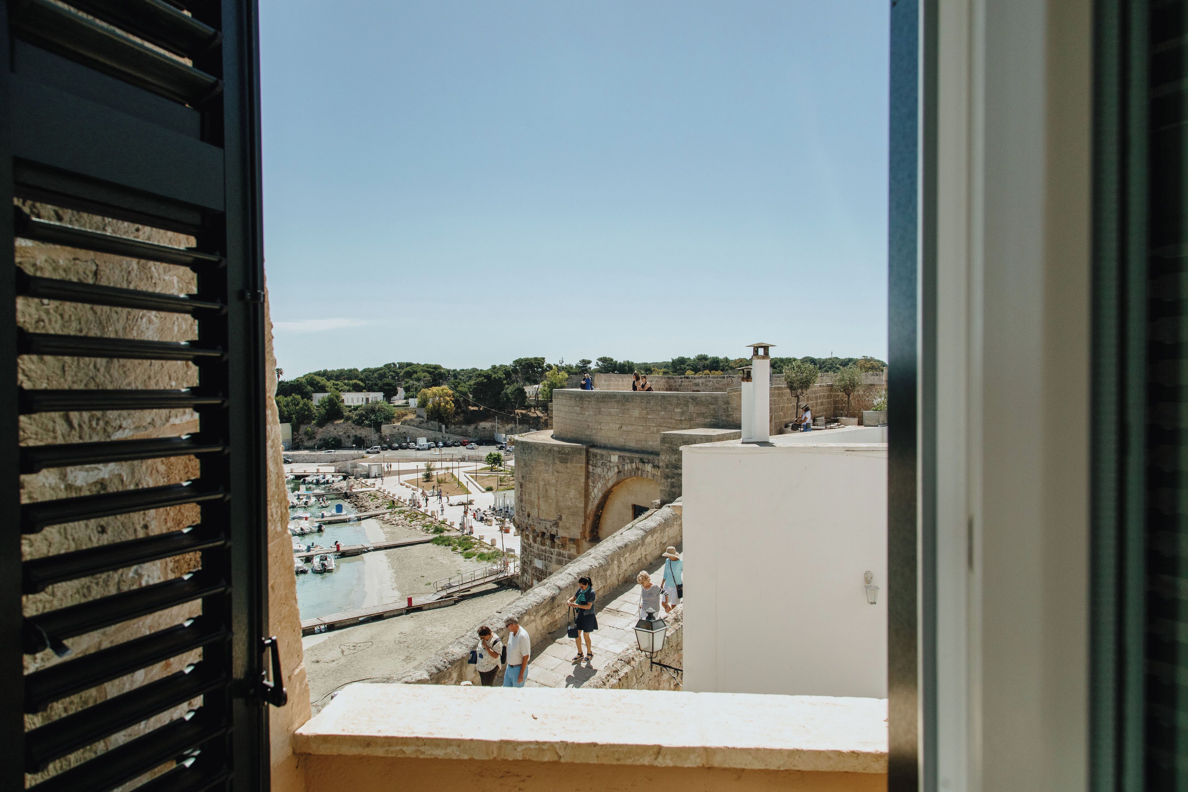 Vista desde una ventana sobre una zona costera con playa, un muro de contención marítimo y un edificio histórico de piedra.