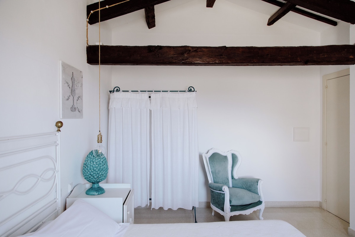 Un dormitorio con una cama blanca, una mesita de noche blanca, una lámpara verde aguamarina, una cortina blanca y una silla verde aguamarina.