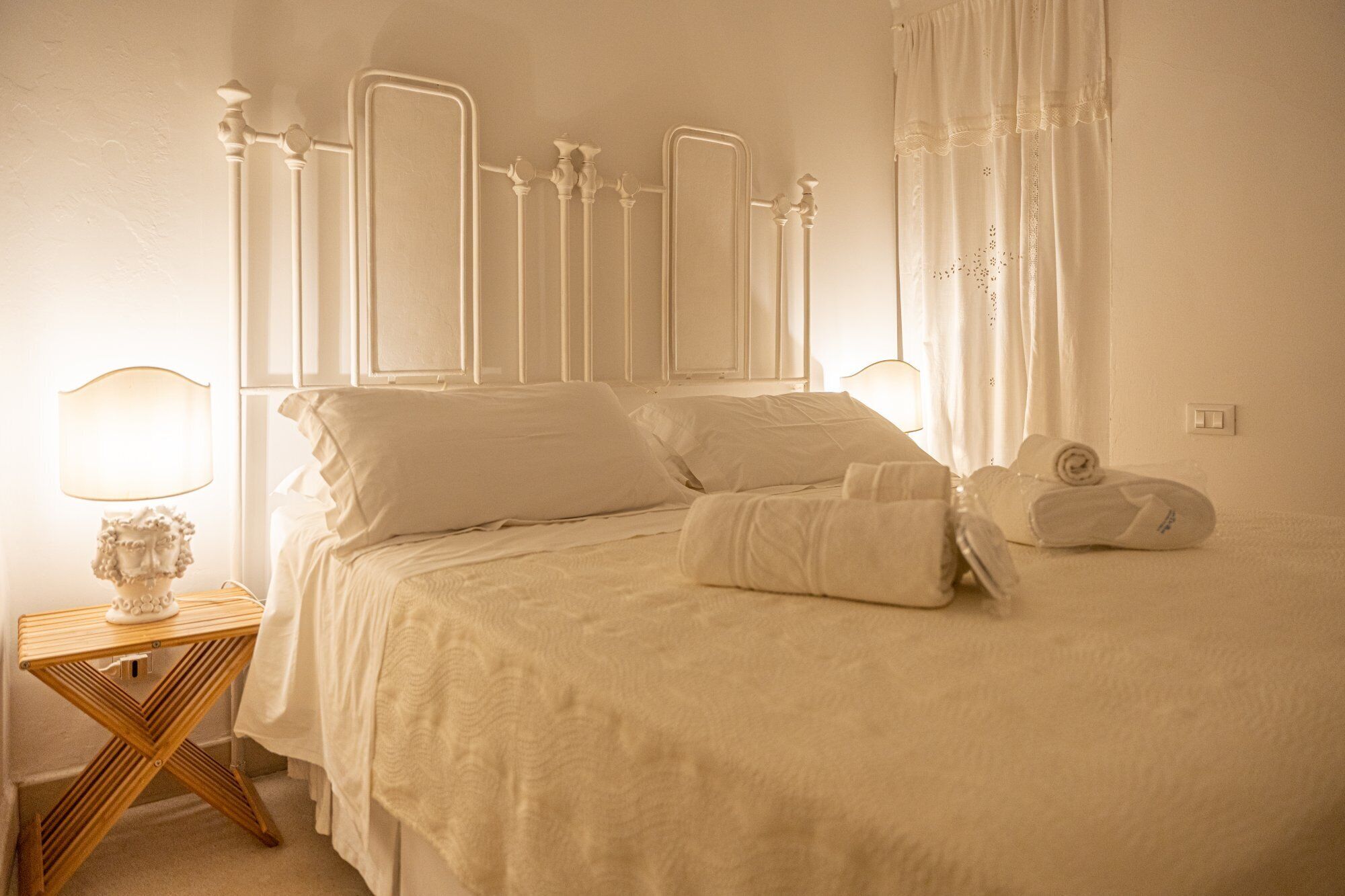 A neatly made bed with white linens and pillows, a wooden nightstand with a lamp, and a white curtain.