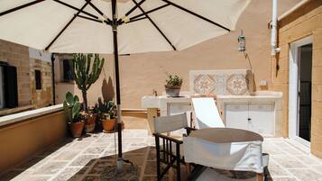 A patio with a table and chairs, a large umbrella, potted plants, and a tiled backsplash.