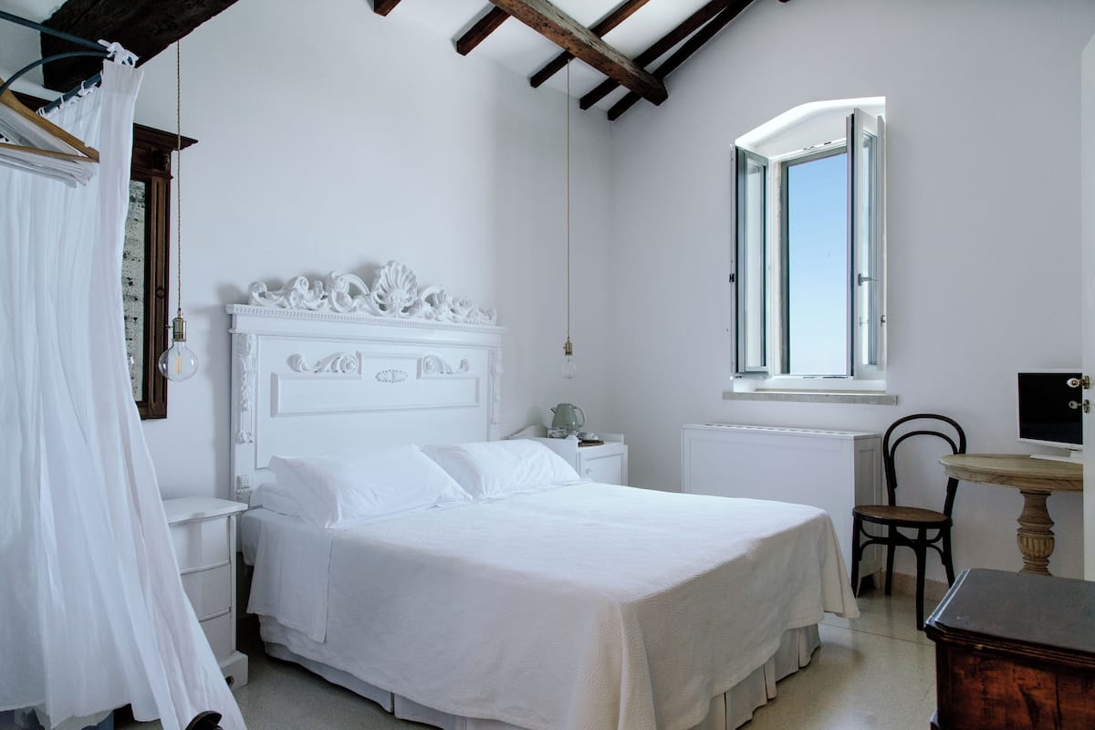 A bedroom with a white bed, a wooden chair, a small table, and a window with daylight.