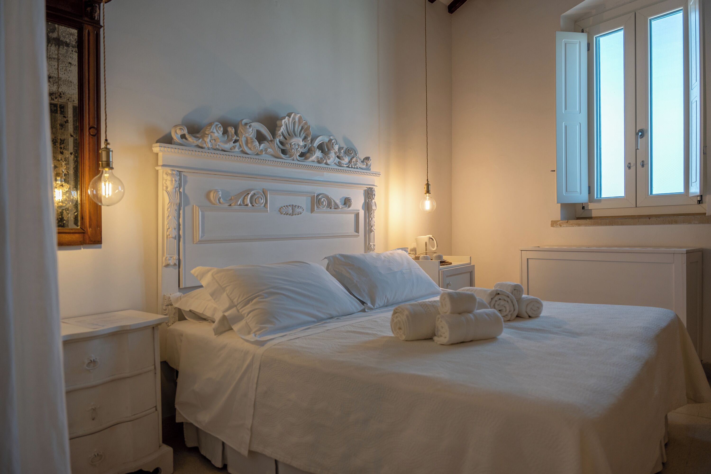 Un lit bien fait, avec du linge de lit blanc et des serviettes pliées, une tête de lit en bois ornée de motifs complexes et une table de chevet avec une lampe.