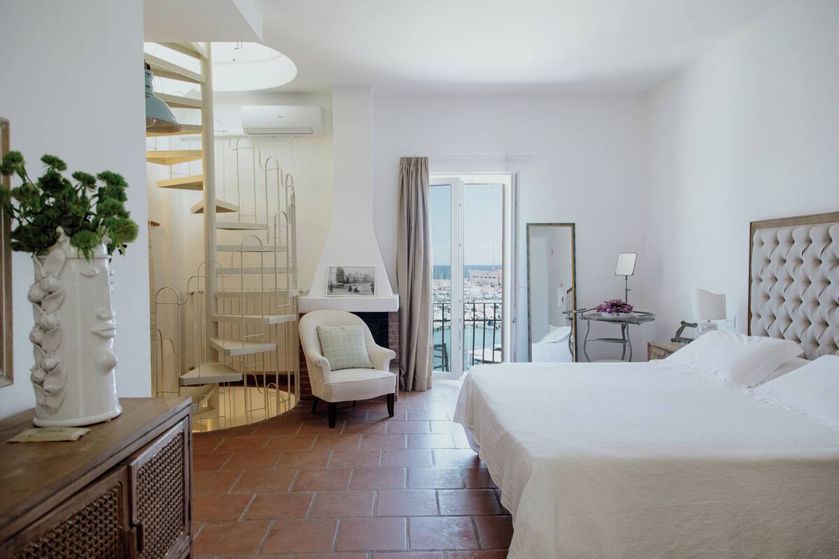 A hotel room with a bed, a chair, a small table, a balcony with a view, and a staircase.