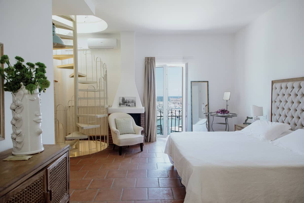A hotel room with a bed, a chair, a small table, a balcony with a view, and a staircase.