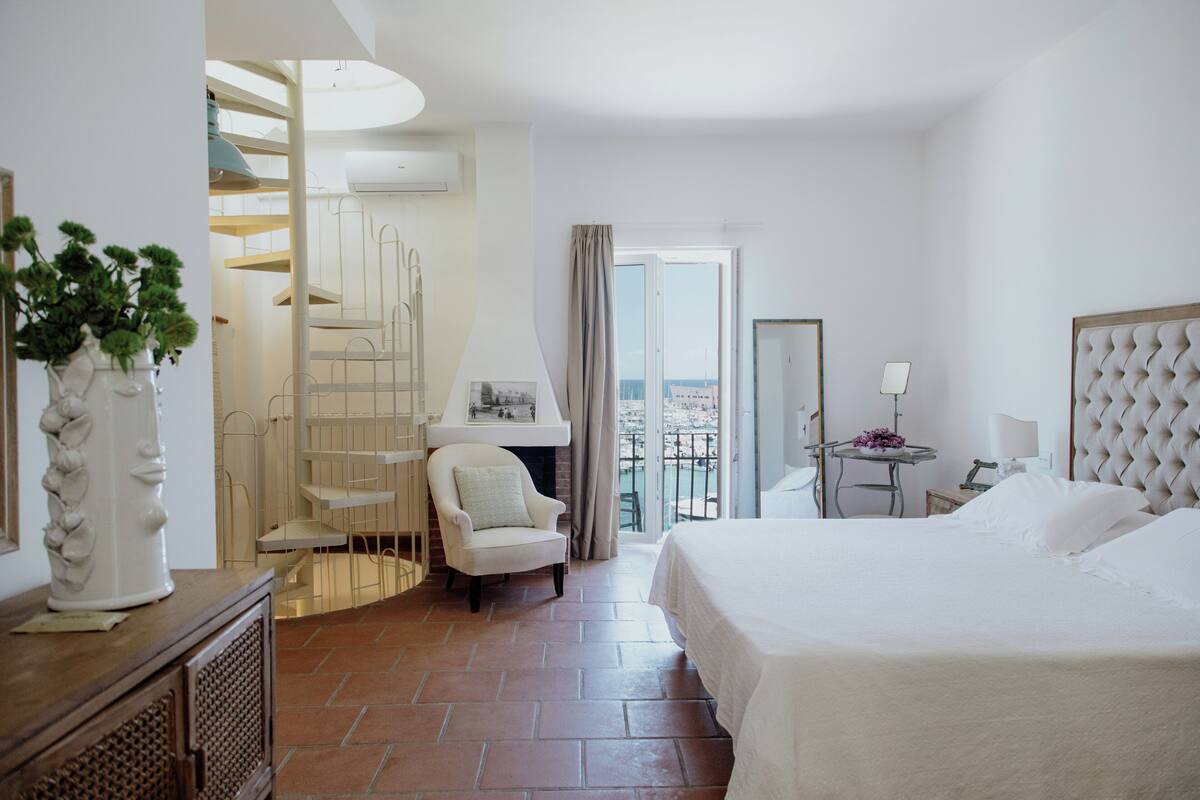 A hotel room with a bed, a chair, a small table, a balcony with a view, and a staircase.