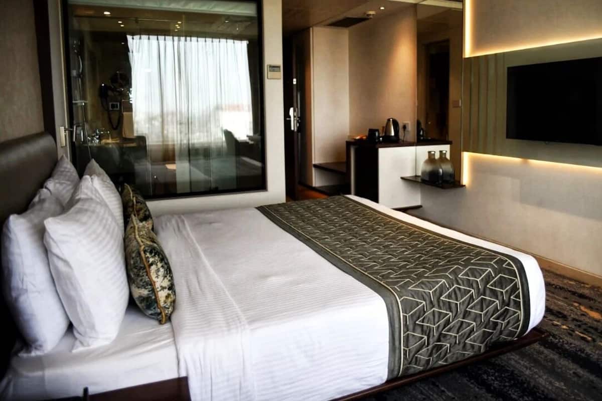 A hotel room with a large bed, a TV mounted on the wall, a desk with a chair, and a glass partition separating the room from the hallway.