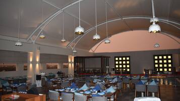 A spacious restaurant with high ceilings and hanging pendant lights. Tables are arranged in a U-shape, and there are large windows providing views of the cityscape.