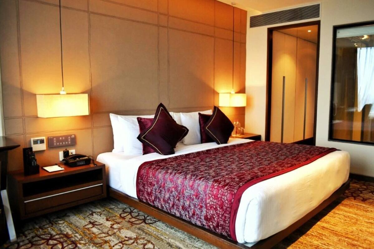 A hotel room with a large bed, two bedside lamps, a nightstand with a phone and a remote, and a window with curtains.