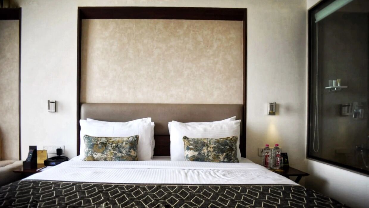 A hotel room with a large bed, two decorative pillows, a headboard, a nightstand with bottles, and a window with a view.