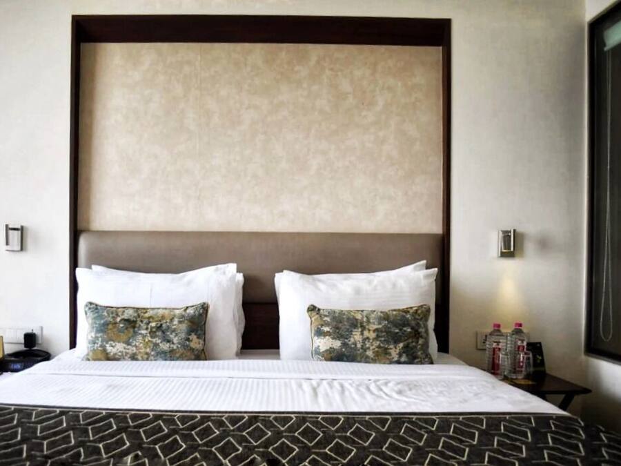 A hotel room with a large bed, two decorative pillows, a headboard, a nightstand with bottles, and a window with a view.