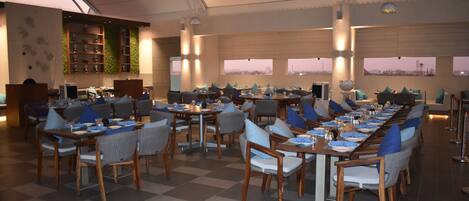 A spacious dining area with multiple tables and chairs, large windows, and a modern ceiling design.