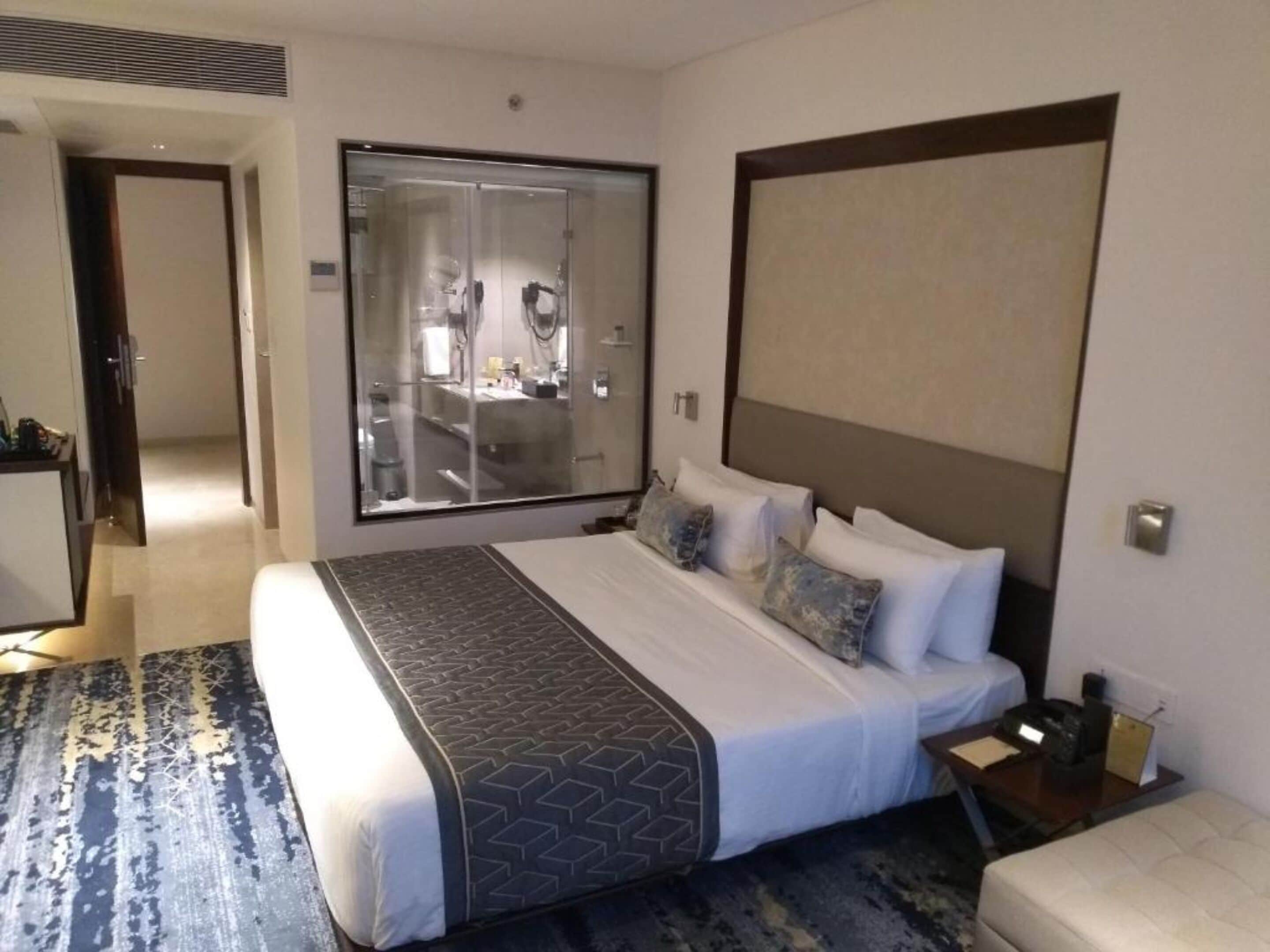 A hotel room with a large bed, a bedside table, a nightstand, a mirror, and a view of the bathroom through a glass door.