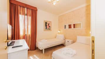 Quarto de hotel com duas camas de solteiro, uma escrivaninha com um laptop e controle remoto, uma mesa de cabeceira com abajur, e uma janela grande com cortinas.
