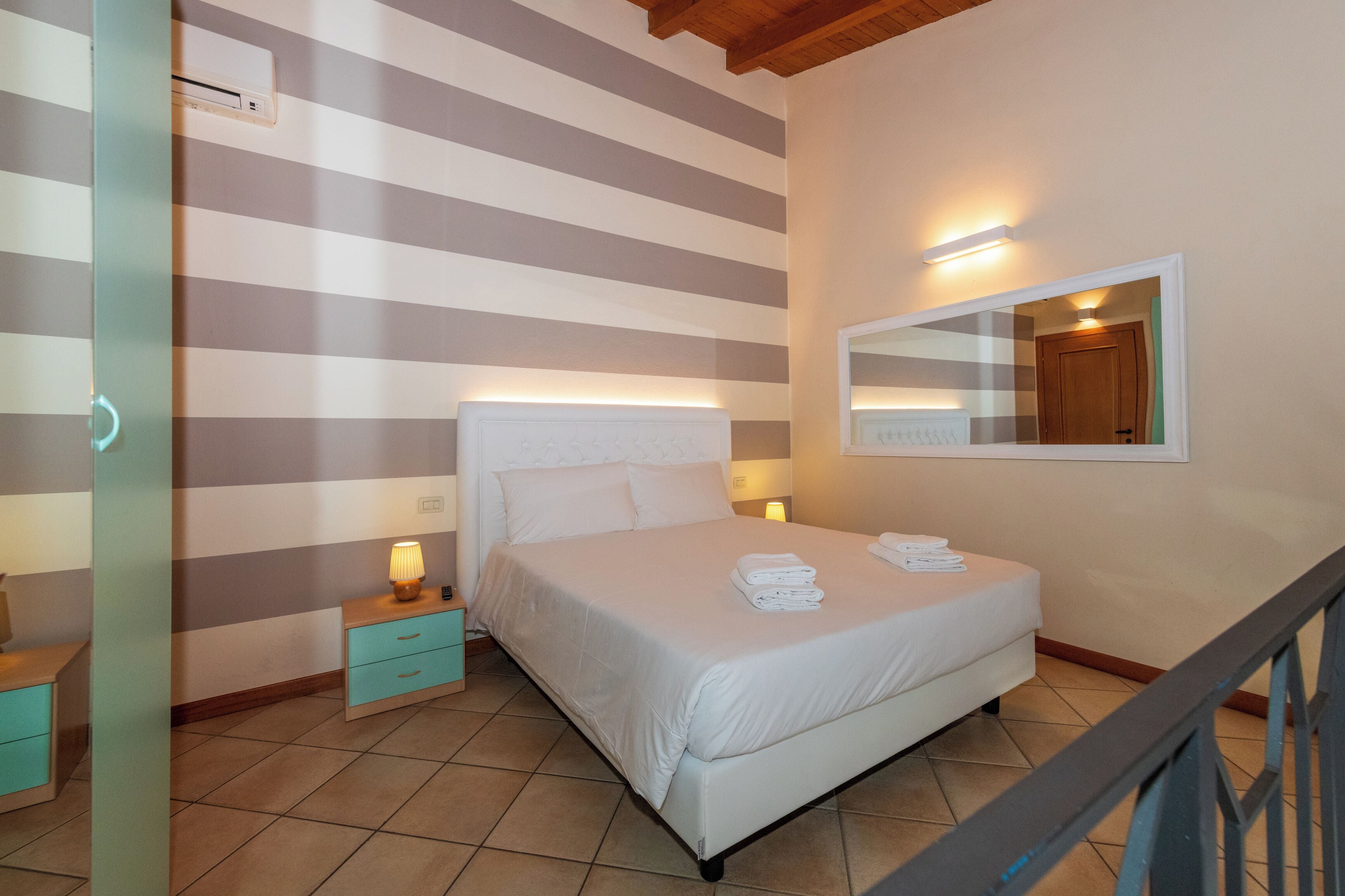 Ein Hotelzimmer mit gestreifter Wand, einem Bett mit weißen Leinen, einem Nachttisch mit Lampe und einem Spiegel an der Wand.