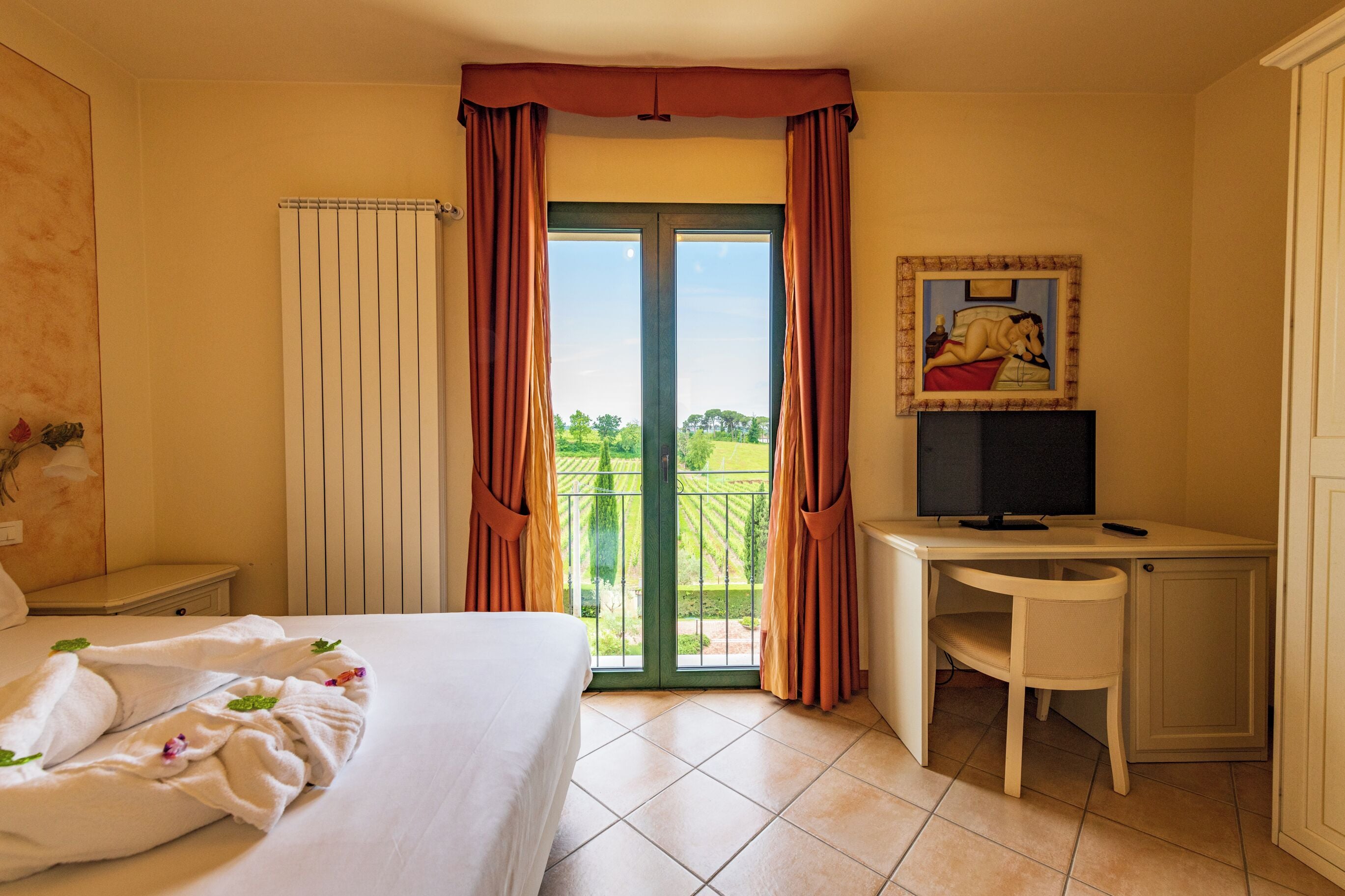 Una camera d'albergo con un letto, una scrivania con un computer, un quadro appeso al muro e un balcone con tende.