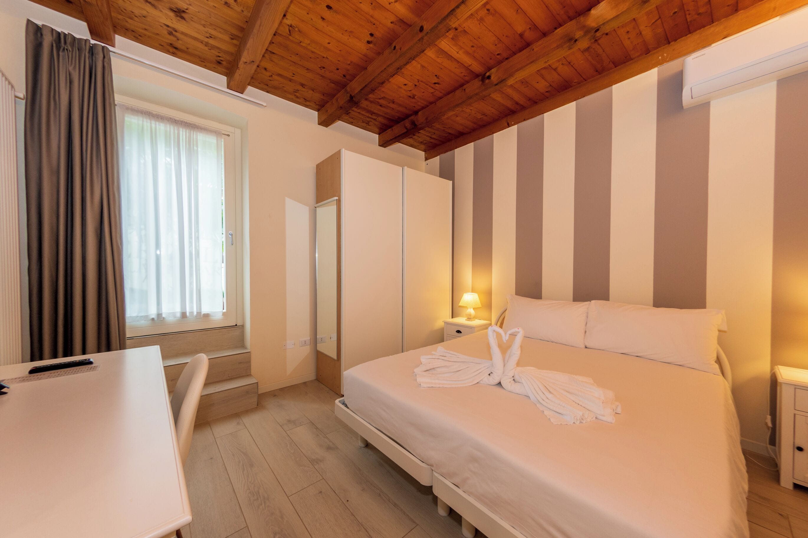 A hotel room with a wooden ceiling, a bed with white bedding, a bedside table with a lamp, and a window with curtains.