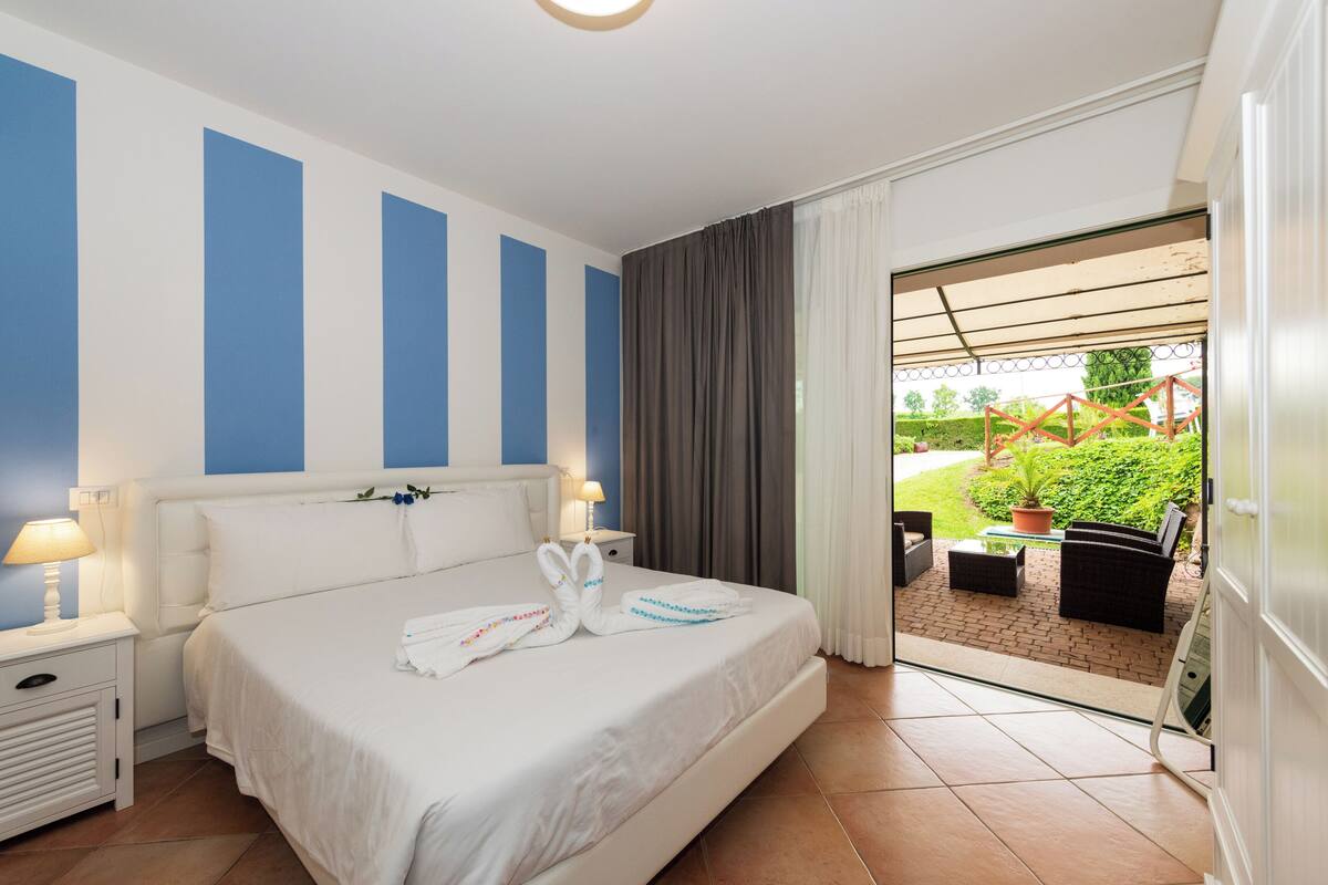 Une chambre d’hôtel avec un grand lit, une table de chevet avec une lampe, une vue sur le jardin depuis la porte, et une petite table avec des chaises à l’extérieur.