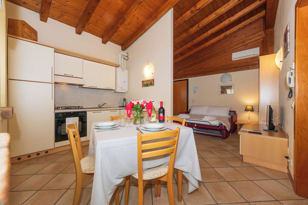 Un coin repas avec une table dressée pour deux personnes, des chaises en bois, une cuisine en arrière-plan et un lit recouvert d’une couverture rouge.