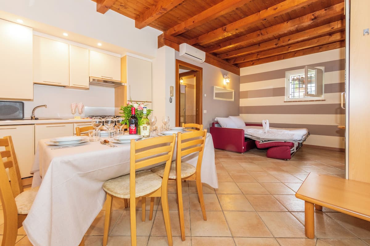 Un coin repas avec une table dressée pour quatre personnes, des chaises en bois et un banc. On aperçoit une petite table avec une chaise rouge à l’arrière-plan.