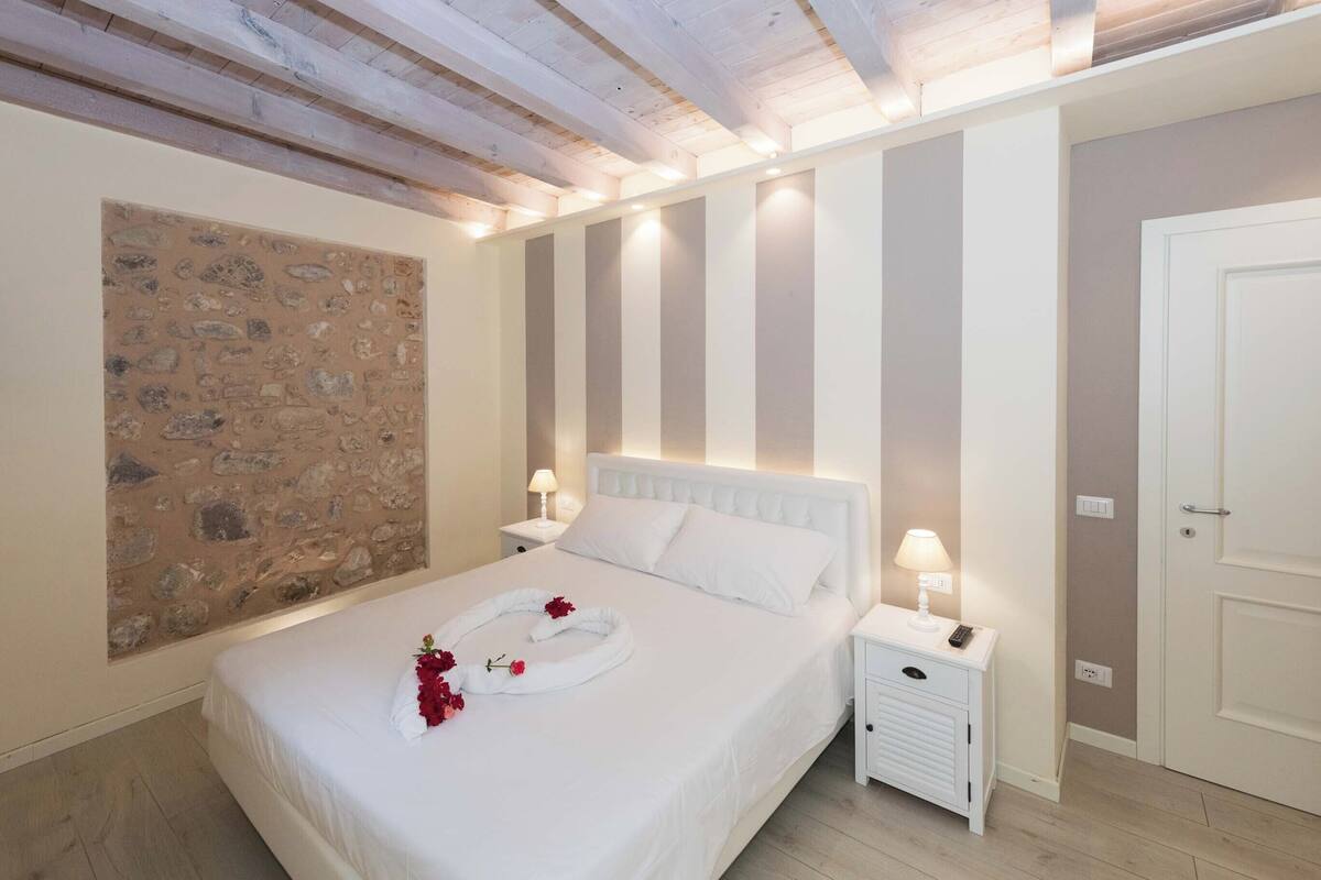 Une chambre à coucher bien aménagée, avec un mur en pierre, un lit recouvert de draps blancs et une table de chevet sur laquelle se trouve une lampe.