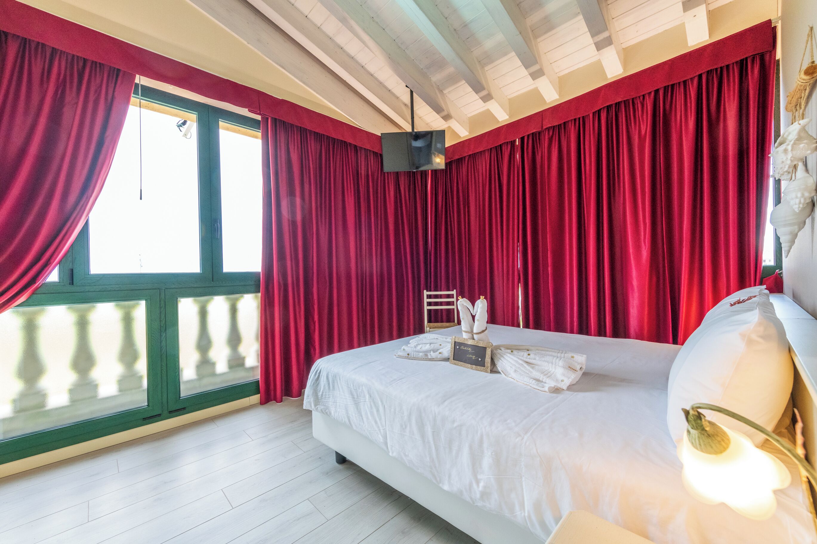 A bedroom with a large bed, red curtains, a balcony with a railing, and a ceiling with exposed beams.