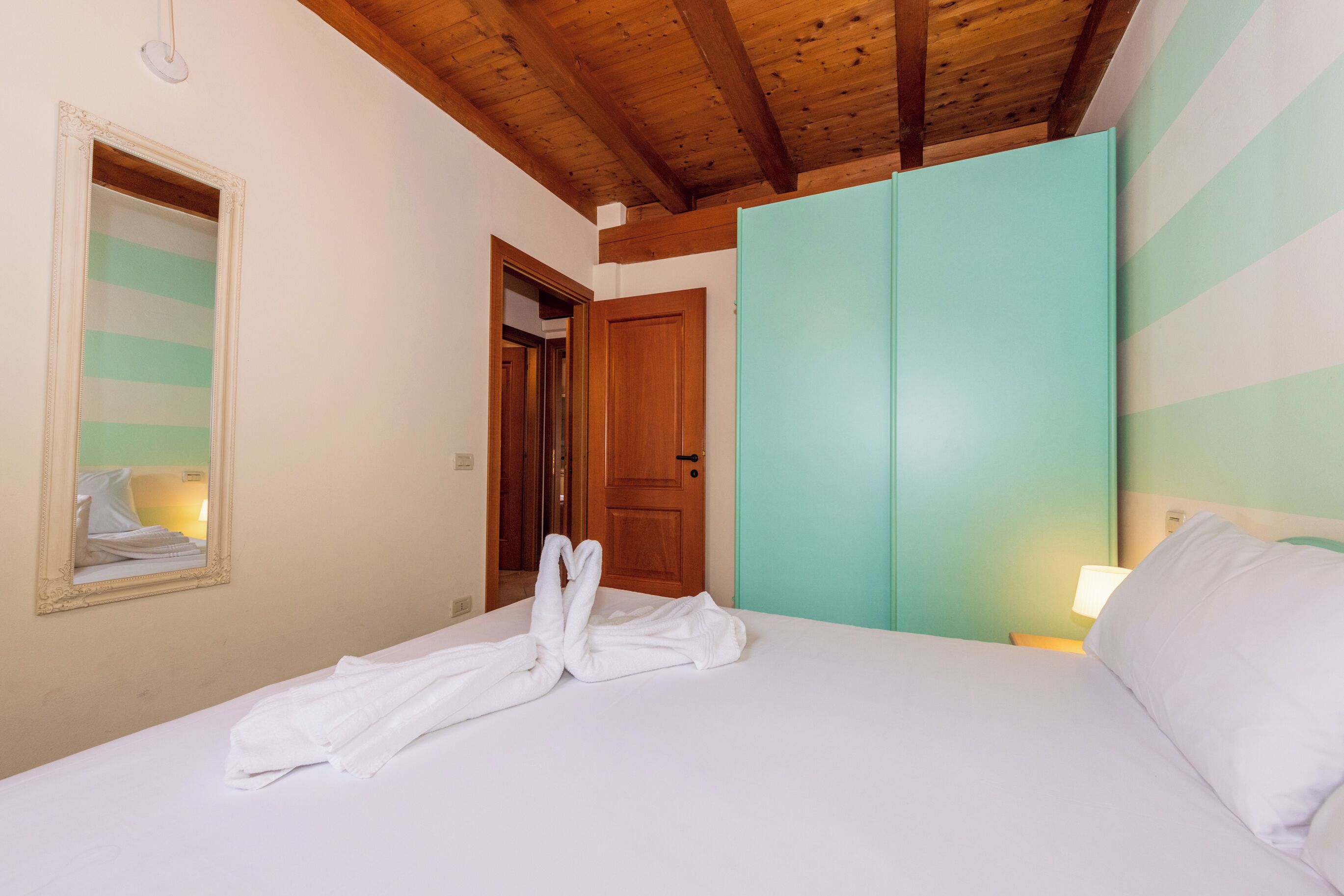 Una camera d'hotel con soffitto in legno, un letto con lenzuola bianche, uno specchio, un armadio color petrolio e un comodino con una lampada.