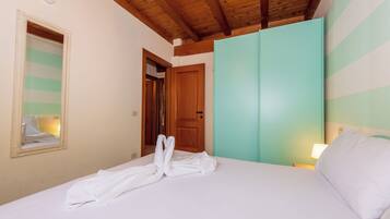 Una camera d'hotel con soffitto in legno, un letto con lenzuola bianche, uno specchio, un armadio color petrolio e un comodino con una lampada.