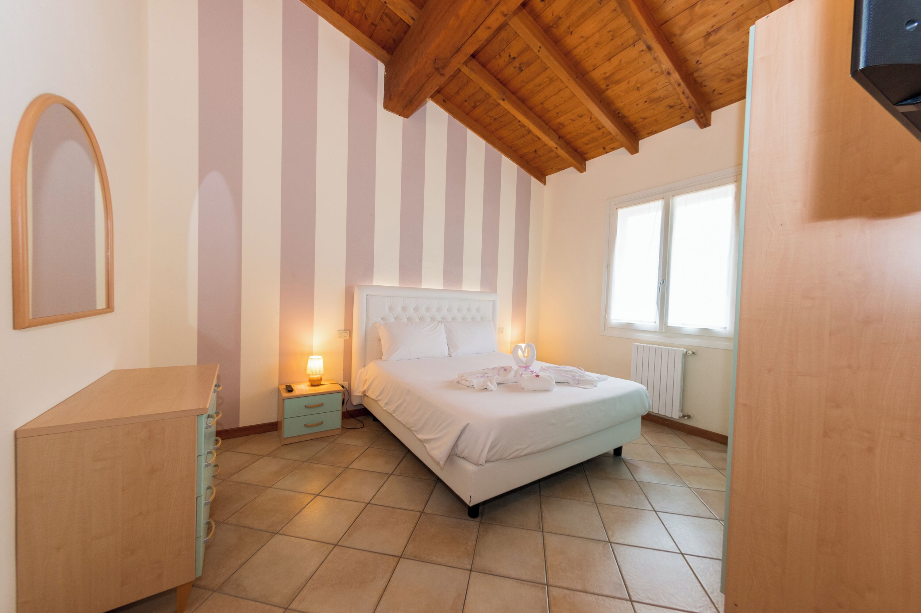 Ein Schlafzimmer mit einem Bett, einem Nachttisch, einer Kommode, einem Fenster und einer Holzbalkendecke.