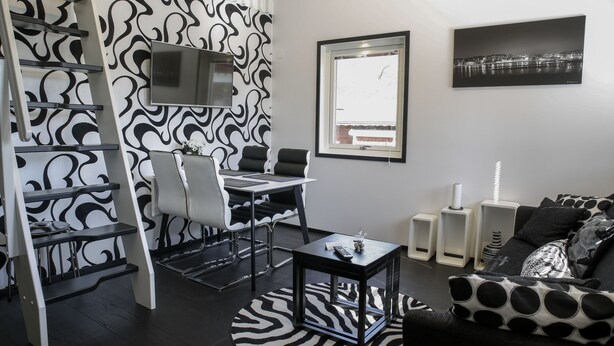 A modern living room with a black and white patterned wall, a flat-screen TV, a cityscape painting, and a cozy seating area with a patterned sofa. - Dragsö Camping & Stugby (Karlskrona)