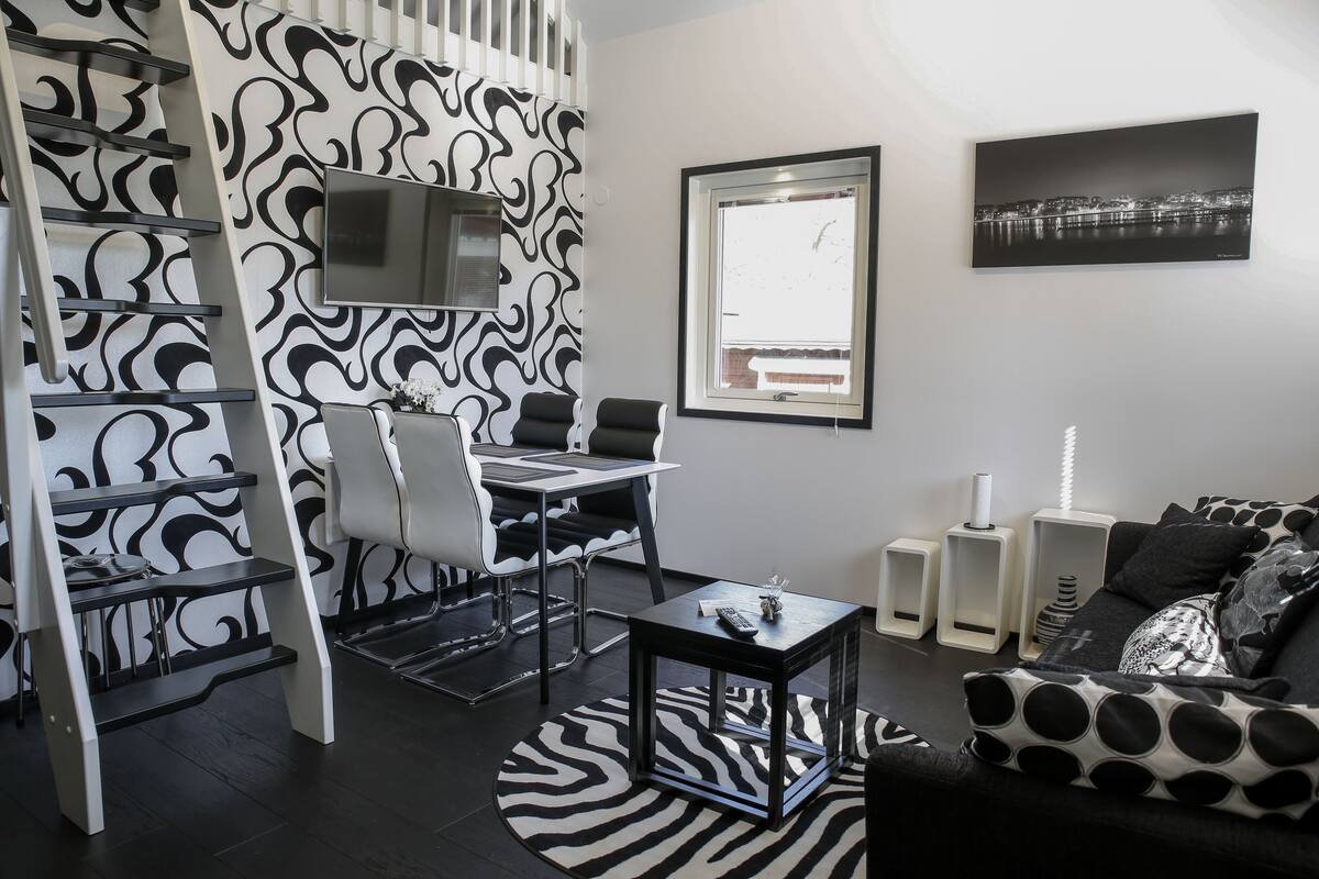 A modern living room with a black and white patterned wall, a flat-screen TV, a cityscape painting, and a cozy seating area with a patterned sofa.